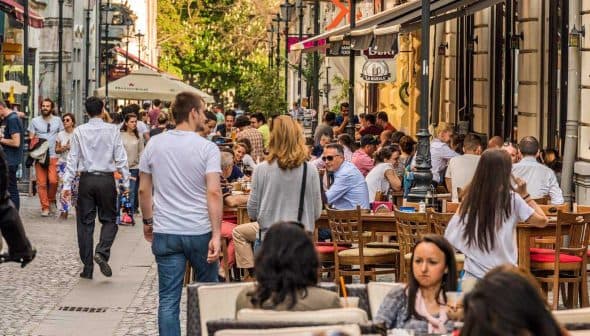 Crowd of people dining at outdoor tables along a city street with pedestrians walking by. - Olive Oil Times
