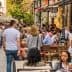 Crowd of people dining at outdoor tables along a city street with pedestrians walking by. - Olive Oil Times