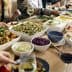 A dining table filled with various dishes, including salads, meats, and side dishes, with people dining around it. - Olive Oil Times