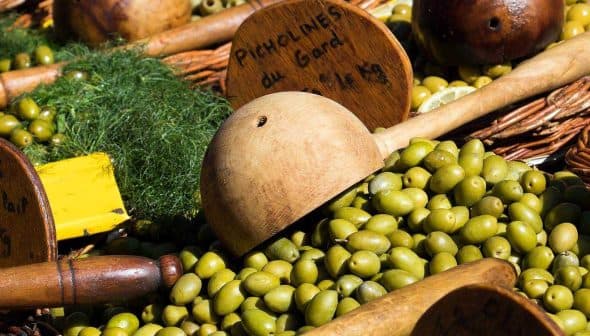 A collection of green olives displayed with wooden utensils and signs indicating prices. - Olive Oil Times