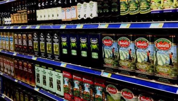 A supermarket shelf displaying various brands of olive oil in bottles and cartons. - Olive Oil Times