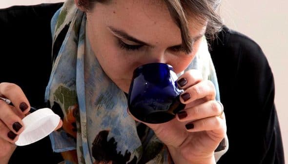 Woman with a scarf smelling from a blue cup while holding a white lid and a pen. - Olive Oil Times