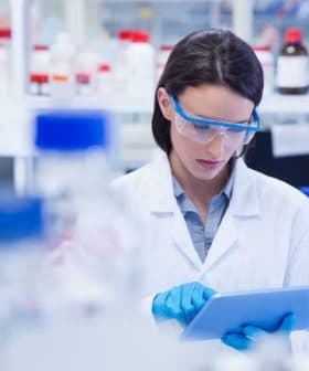 Female scientist wearing a lab coat and safety goggles using a tablet in a laboratory setting. - Olive Oil Times
