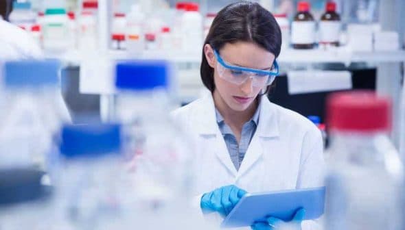 Female scientist wearing a lab coat and safety goggles using a tablet in a laboratory setting. - Olive Oil Times