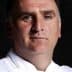 Portrait of a chef wearing a white uniform with a serious expression against a dark background. - Olive Oil Times