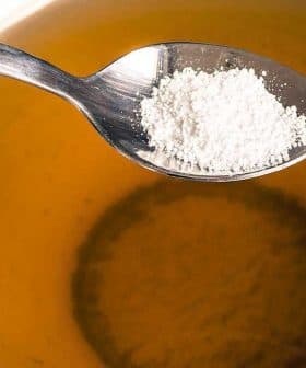 A spoon holding sugar above a cup of tea filled with a light brown liquid. - Olive Oil Times