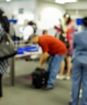 Blurred image of passengers and airport staff in a check-in area with luggage and service counters. - Olive Oil Times