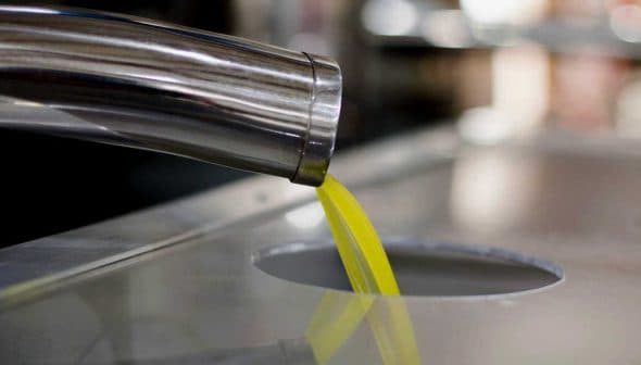 Stream of olive oil pouring from a metal dispenser into a circular opening. - Olive Oil Times