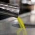 Stream of olive oil pouring from a metal dispenser into a circular opening. - Olive Oil Times