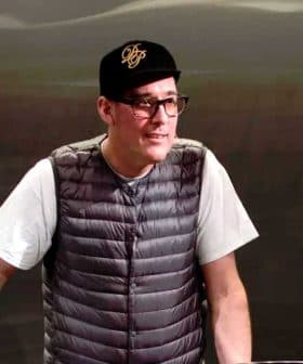 Man wearing a gray vest and black cap standing at a table during a presentation. - Olive Oil Times
