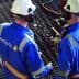 Two Petrofac workers wearing blue uniforms and safety helmets discussing on a metal grid surface. - Olive Oil Times