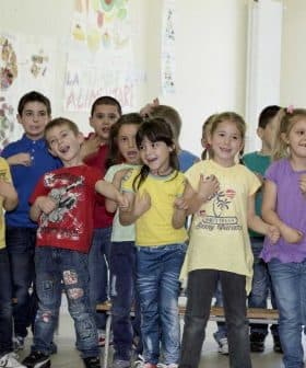 A group of children in colorful shirts singing together in a classroom setting. - Olive Oil Times