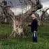 A man in a black jacket standing next to an olive tree in a field. - Olive Oil Times