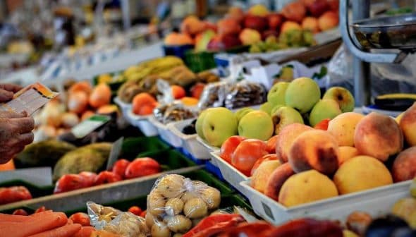 A selection of fresh fruits and vegetables displayed at a market stall, with a person holding a shopping list. - Olive Oil Times