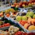 A selection of fresh fruits and vegetables displayed at a market stall, with a person holding a shopping list. - Olive Oil Times