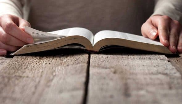 Hands turning pages of an open book resting on a wooden table. - Olive Oil Times