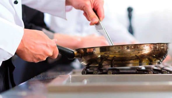 Chef in a white shirt using a frying pan on a gas stove in a kitchen setting. - Olive Oil Times