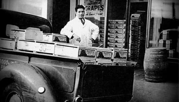 A man in a white coat standing beside a vintage grocery delivery truck filled with boxes of grapes. - Olive Oil Times