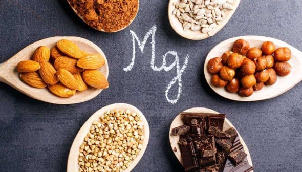 Wooden spoons containing almonds, hazelnuts, sunflower seeds, and cocoa powder arranged around the letter 'Mg' written in chalk. - Olive Oil Times
