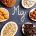 Wooden spoons containing almonds, hazelnuts, sunflower seeds, and cocoa powder arranged around the letter 'Mg' written in chalk. - Olive Oil Times