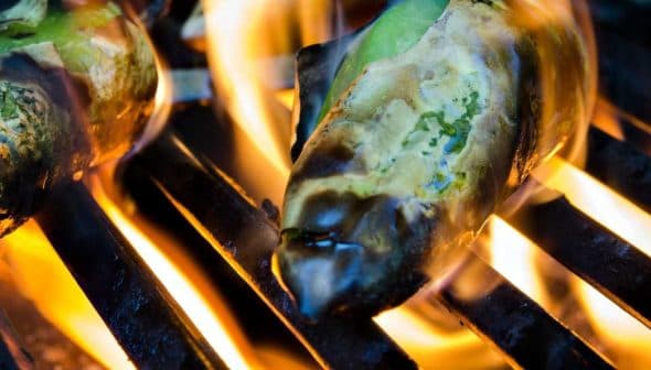 Green peppers being grilled on a barbecue grill with flames visible. - Olive Oil Times