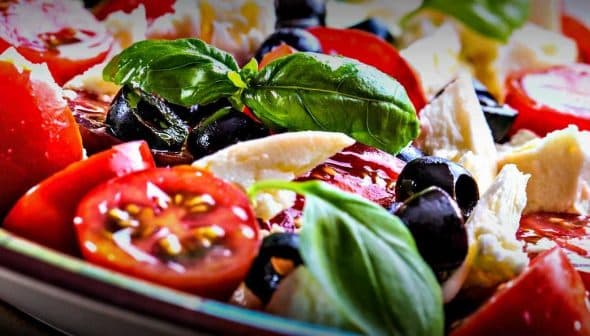 A bowl of mixed salad featuring cherry tomatoes, black olives, and fresh basil leaves. - Olive Oil Times