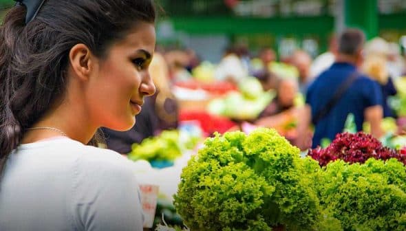 A woman looking at fresh vegetables in a market, with various greens and produce visible in the background. - Olive Oil Times
