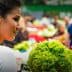 A woman looking at fresh vegetables in a market, with various greens and produce visible in the background. - Olive Oil Times
