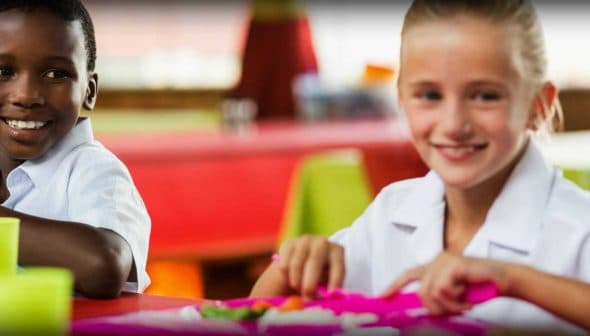 Two children smiling while eating lunch at a colorful table in a dining area. - Olive Oil Times