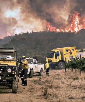 Firefighters and vehicles responding to a wildfire in a rural area with smoke and flames in the background. - Olive Oil Times