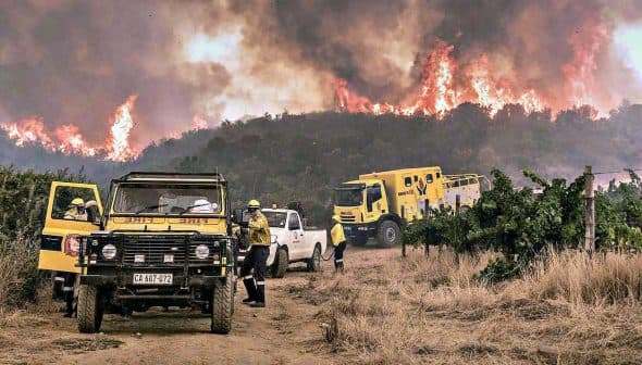 Firefighters and vehicles responding to a wildfire in a rural area with smoke and flames in the background. - Olive Oil Times