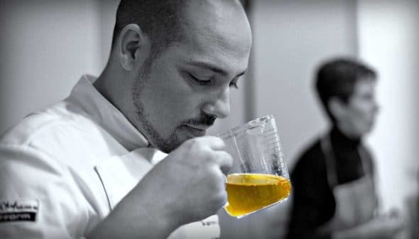 Chef smelling olive oil from a measuring cup while another person is in the background. - Olive Oil Times