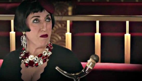 Woman seated at a restaurant wearing a black dress and a large statement necklace with red and silver accents. - Olive Oil Times