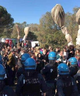 Police officers in riot gear facing a crowd at an outdoor event with large fabric banners. - Olive Oil Times