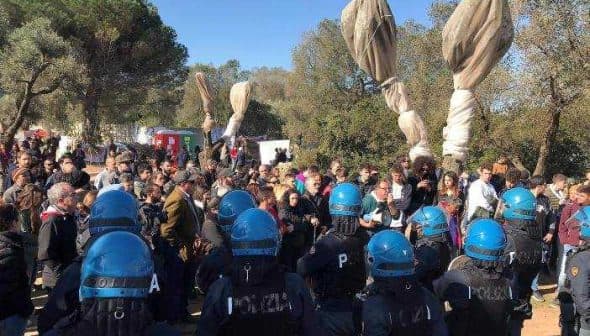Police officers in riot gear facing a crowd at an outdoor event with large fabric banners. - Olive Oil Times