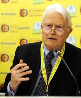 An elderly man with glasses speaking at a conference table with a yellow background. - Olive Oil Times