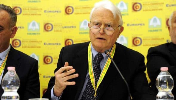 An elderly man with glasses speaking at a conference table with a yellow background. - Olive Oil Times