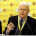 An elderly man with glasses speaking at a conference table with a yellow background. - Olive Oil Times