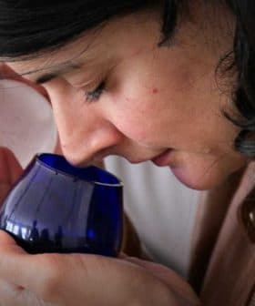 Woman with dark hair holding a blue glass container close to her face, smelling the contents. - Olive Oil Times