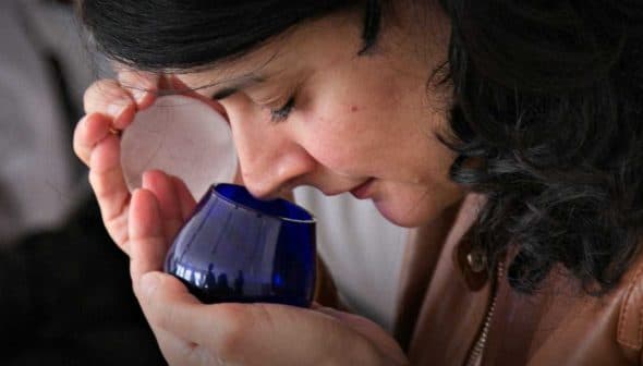 Woman with dark hair holding a blue glass container close to her face, smelling the contents. - Olive Oil Times