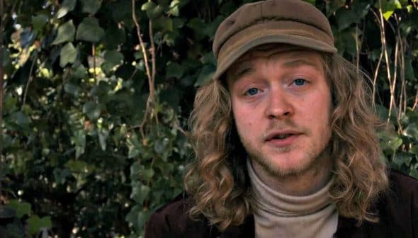 A man with long, curly hair wearing a beige cap and a turtleneck sweater, sitting outdoors. - Olive Oil Times