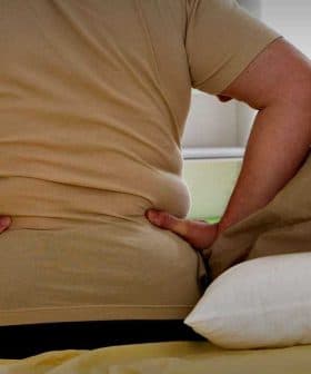 Individual sitting on a bed with hands on lower back, indicating discomfort or pain. - Olive Oil Times
