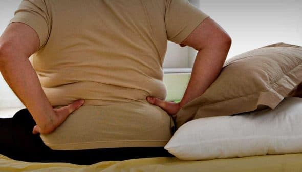 Individual sitting on a bed with hands on lower back, indicating discomfort or pain. - Olive Oil Times