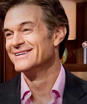 A man with light brown hair smiling while seated in a studio with various objects in the background. - Olive Oil Times