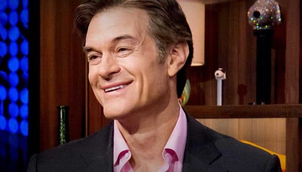 A man with light brown hair smiling while seated in a studio with various objects in the background. - Olive Oil Times