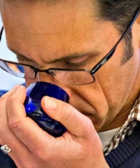 A man wearing glasses and a checkered shirt smelling olive oil from a blue glass cup. - Olive Oil Times
