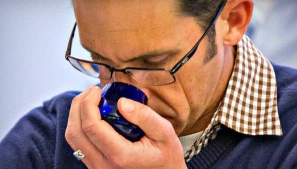 A man wearing glasses and a checkered shirt smelling olive oil from a blue glass cup. - Olive Oil Times