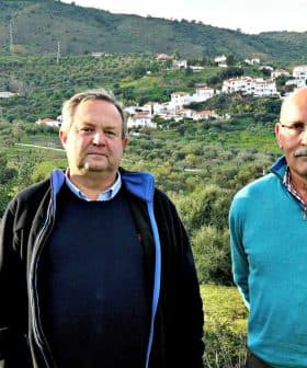 Two men standing side by side in an olive grove with a hillside village in the background. - Olive Oil Times