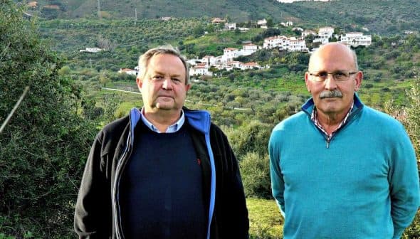 Two men standing side by side in an olive grove with a hillside village in the background. - Olive Oil Times