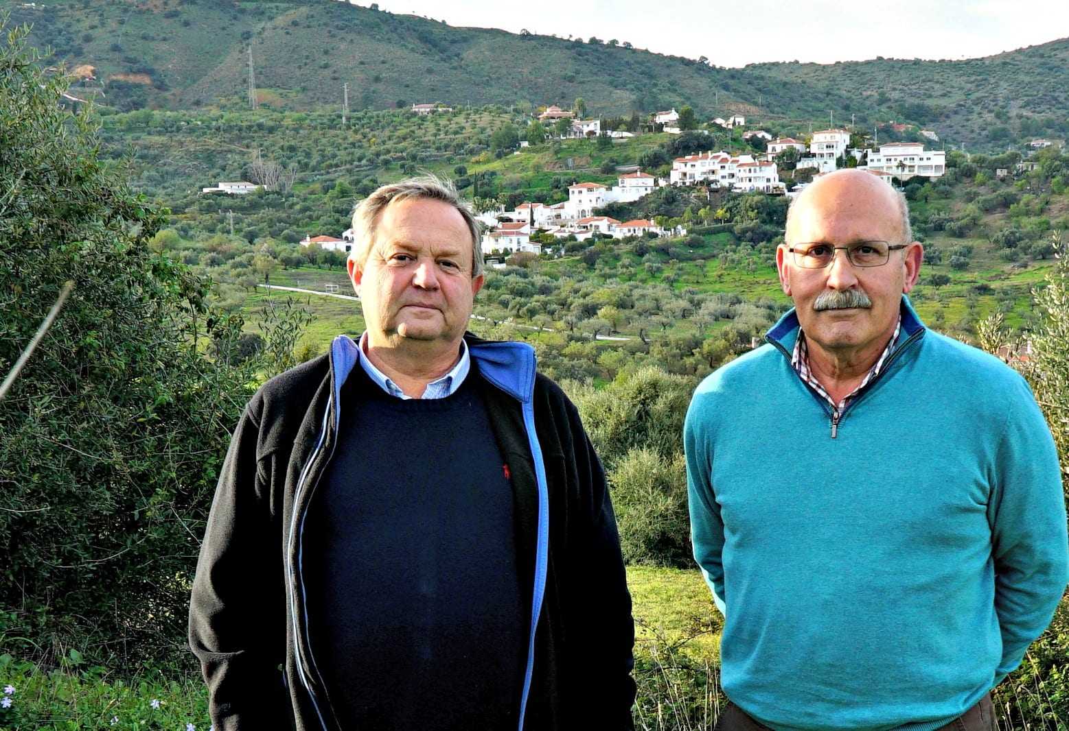 Two men standing side by side in an olive grove with a hillside village in the background. - Olive Oil Times